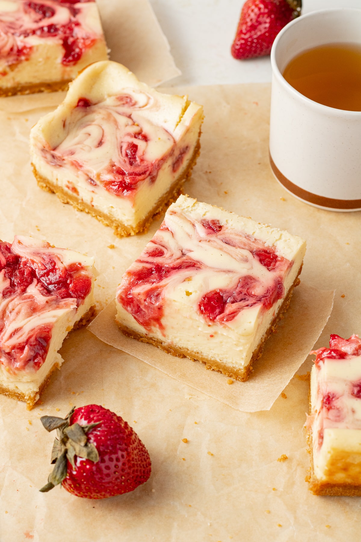 3/4 view of cheesecake bars on pieces of parchment paper with strawberries and a cup of tea near by.
