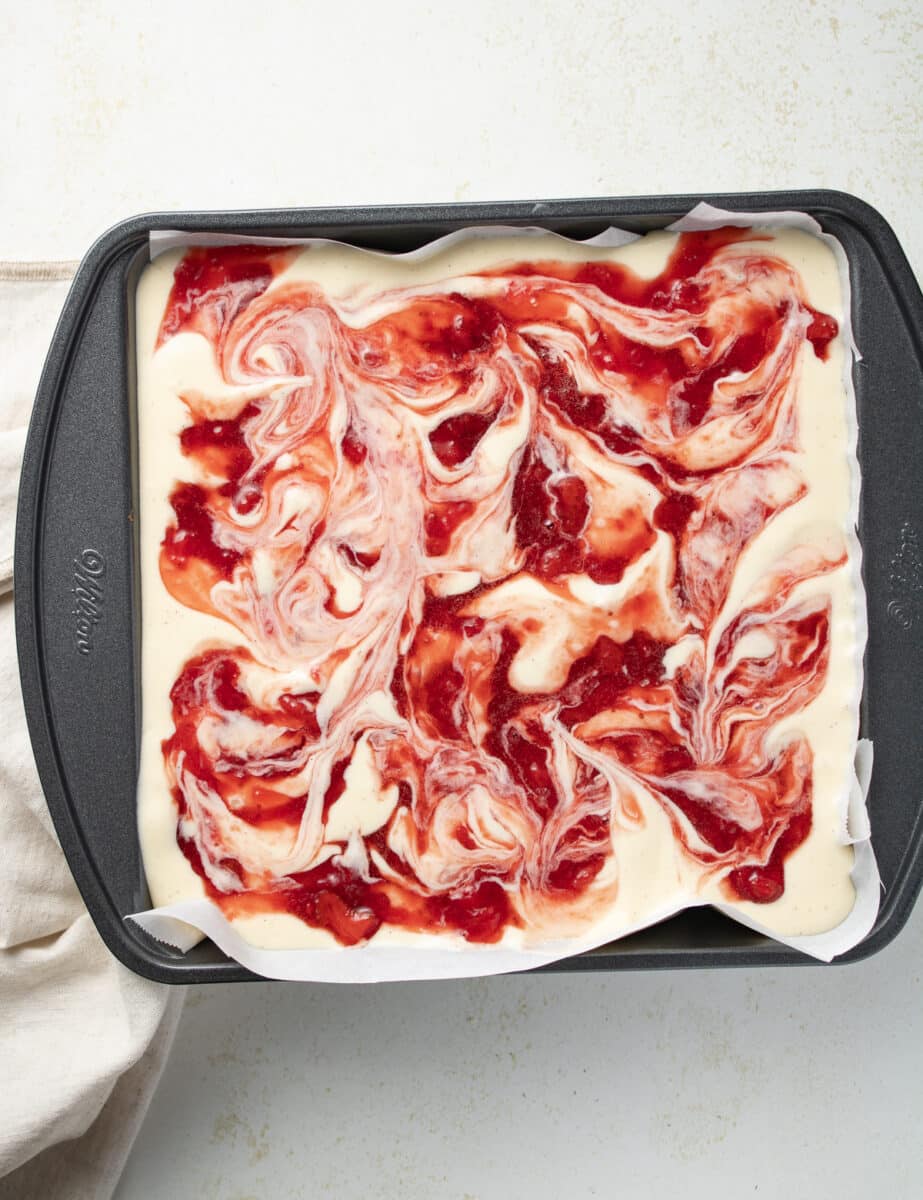 Cheesecake batter with strawberry sauce swirled on top in a cake pan.